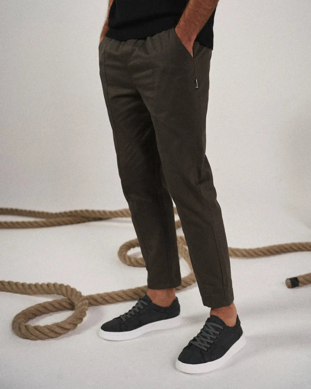 Person wearing dark green pants and black sneakers on a light background with ropes.