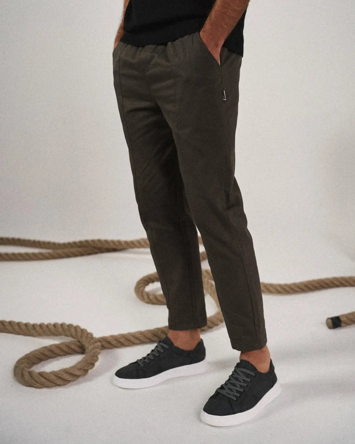 Person wearing dark green pants and black sneakers on a light background with ropes.